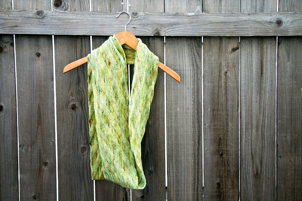 A beautifully textural scarf drapes from a tan wooden hanger resting against a grey wooden fence.
