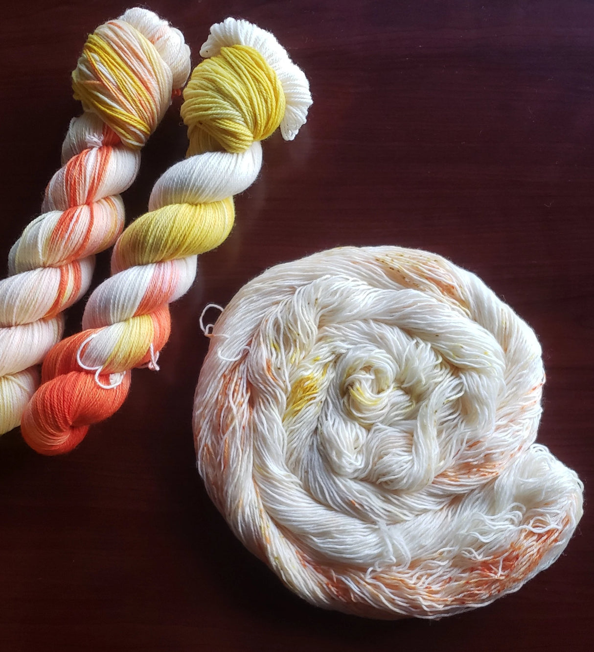 Two skeins of yarn rest in the upper left corner of the frame while a third rests in the mid-bottom right in a curled cake against a dark maroon background.