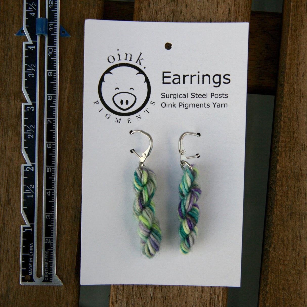 miniature skein earrings rest on a white card with the Oink Pigments logo and text at the top of the card. a small ruler rests on the left.