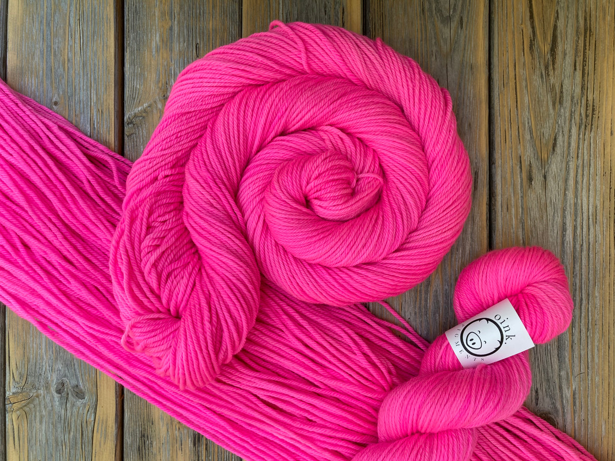 Three skeins of intensely pink yarn rest against a wooden background in various degrees of untwisting; a flat hank, a swirled skein, and a twisted skein with a small white label.
