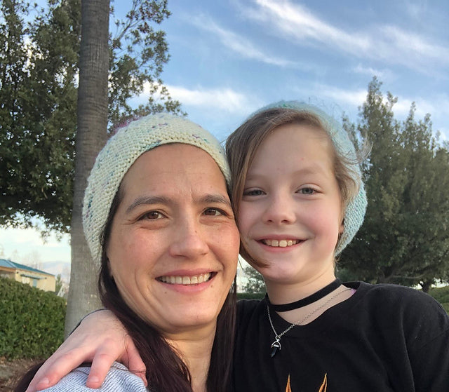 an adult and a child pose for the camera, each wearing knitted hats. They are smiling and have arms around one another.