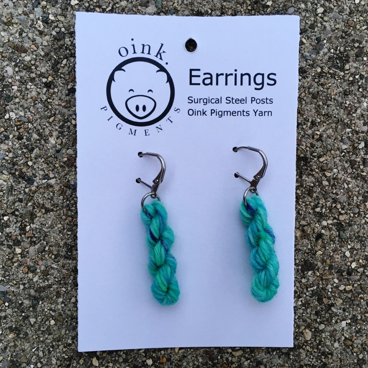 miniature skeins of yarn dangle from steel earring hooks on a white card with the Oink Pigments logo and information at the top.