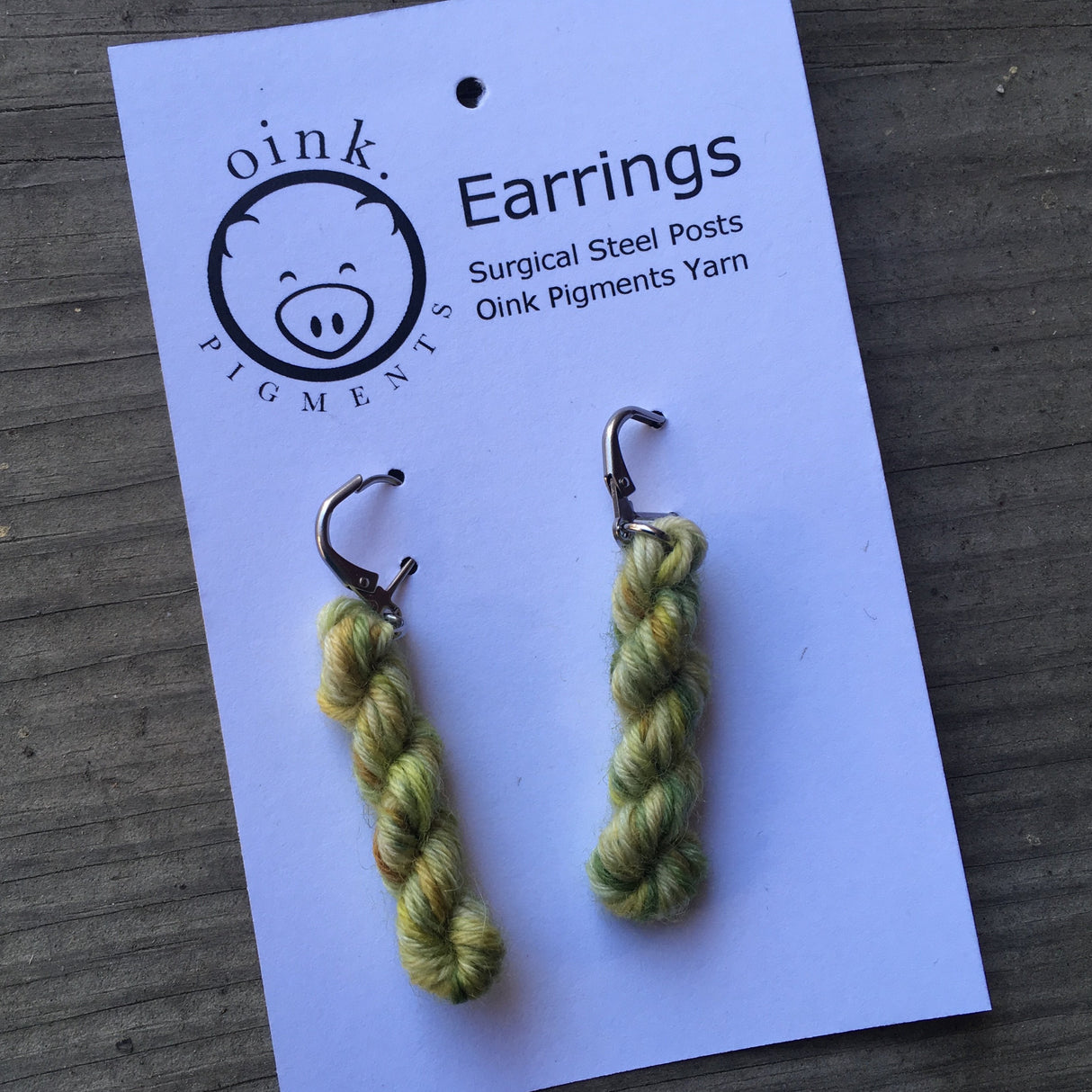 miniature skeins of yarn dangle from steel earring hooks on a white card with the Oink Pigments logo and information at the top.