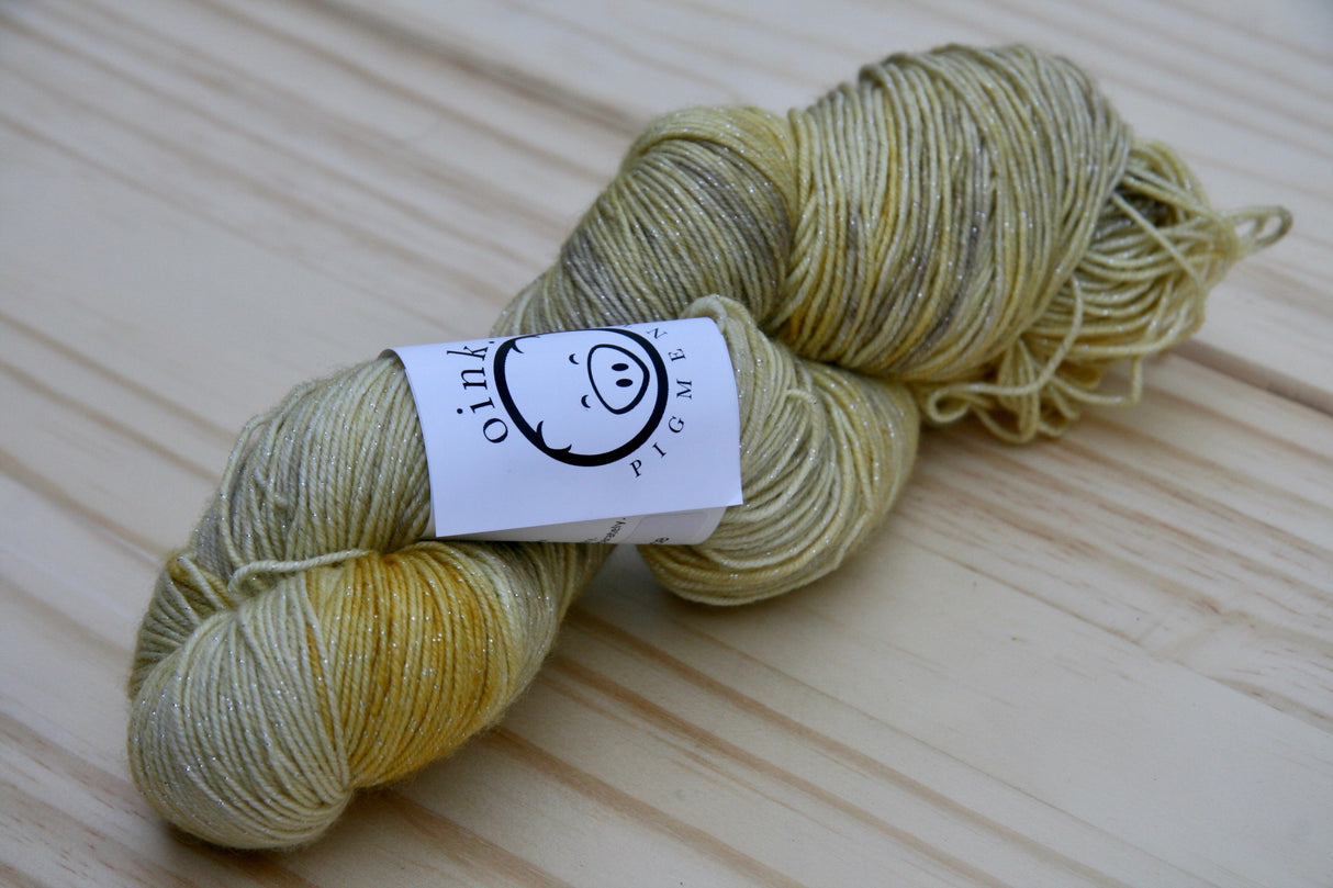Golden tones are highlighted with subtle sparkle. The skein rests on a pale wooden background.