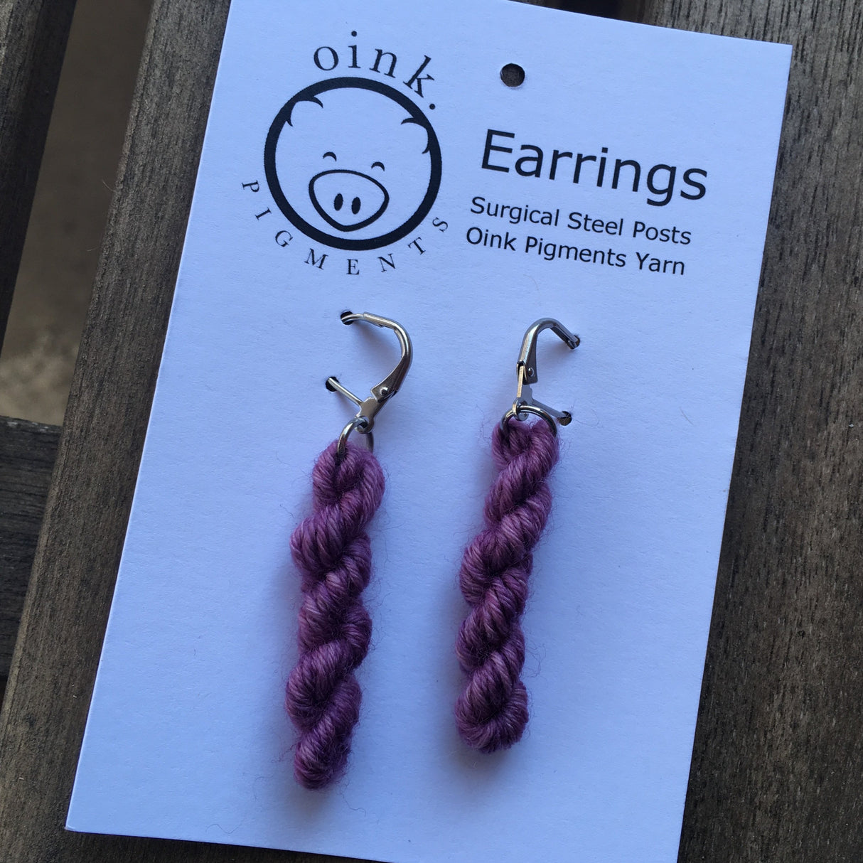 miniature skeins hang from steel ear loops on a white card with the Oink Pigments logo at the top. The background is light wood.