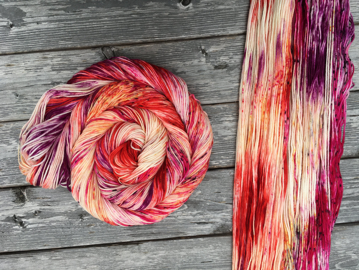 A swirl of yarn rests to the left of a unhanked skein. Both skeins rest on a wooden background.