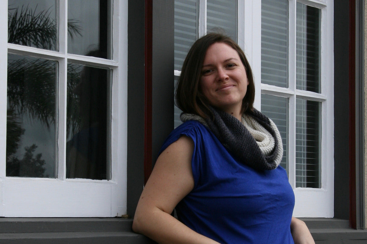 A sweetly smiling person stands in front of windows wearing a royal blue top and a bold, graphic cowl in charcoal grey and cream.