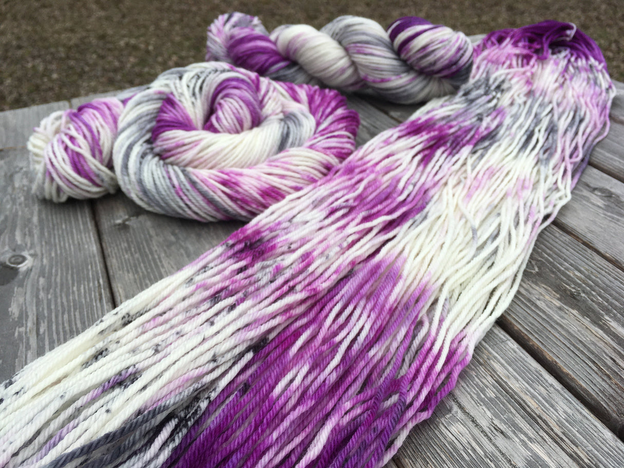 A flat swath of yarn rests diagonally in the foreground of the frame, with a swirled cake and a twist of yarn in the upper background. The yarn is sitting against a grey wooden background.