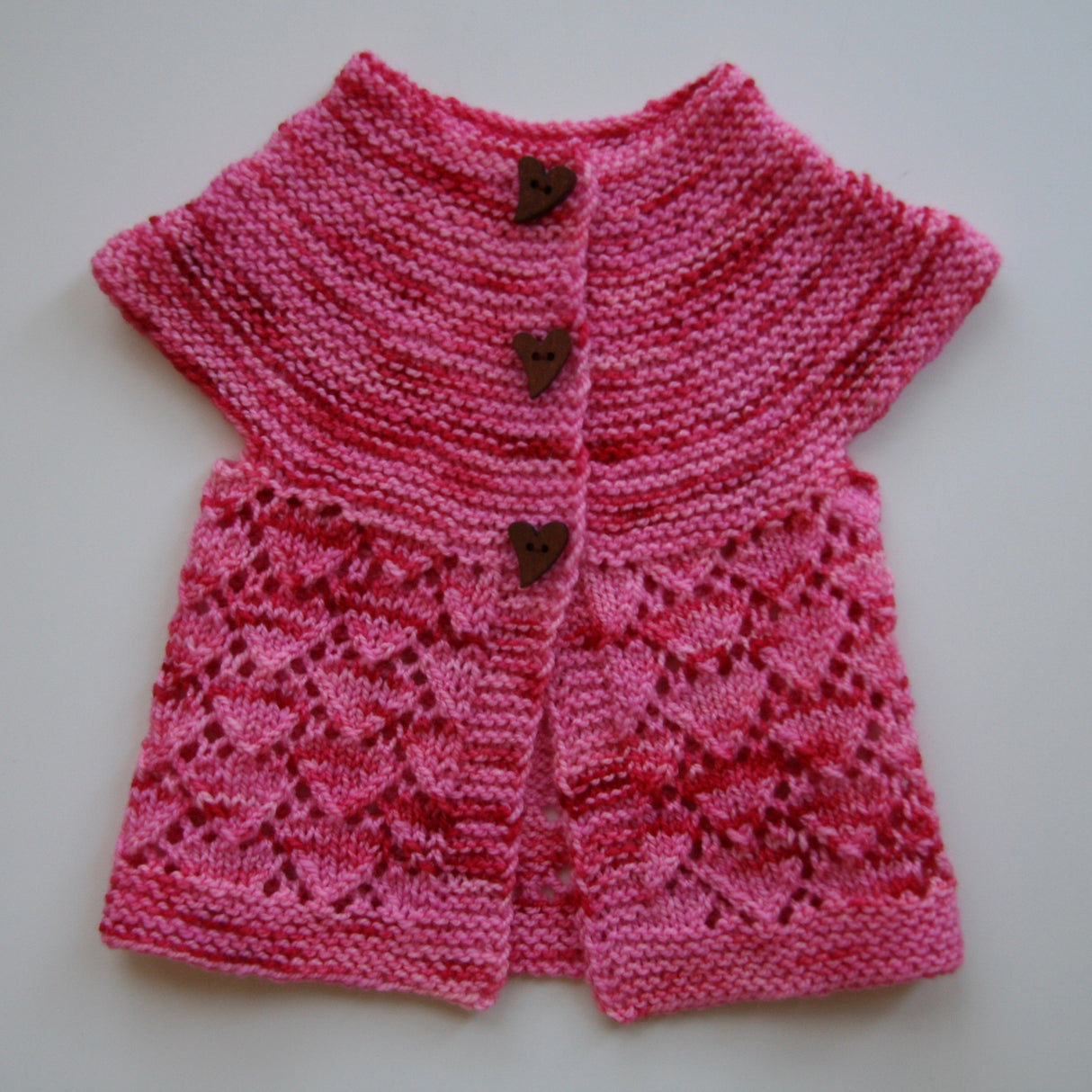 A petite pink and sweater with red streaks sits atop a white background. The top of the little sweater is in a swooping garter stitch, and the body has a delicate lacy heart pattern. Three dark heart buttons complete the darling look.