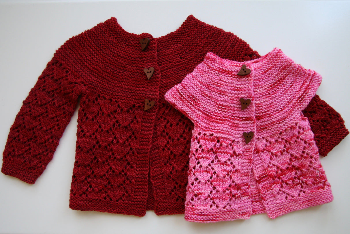 Two sweaters cuddle against a clean white background. The foreground sweater is small and pink, while the background is red and larger. They are both variations on a pattern of heart lace and garter stitch.