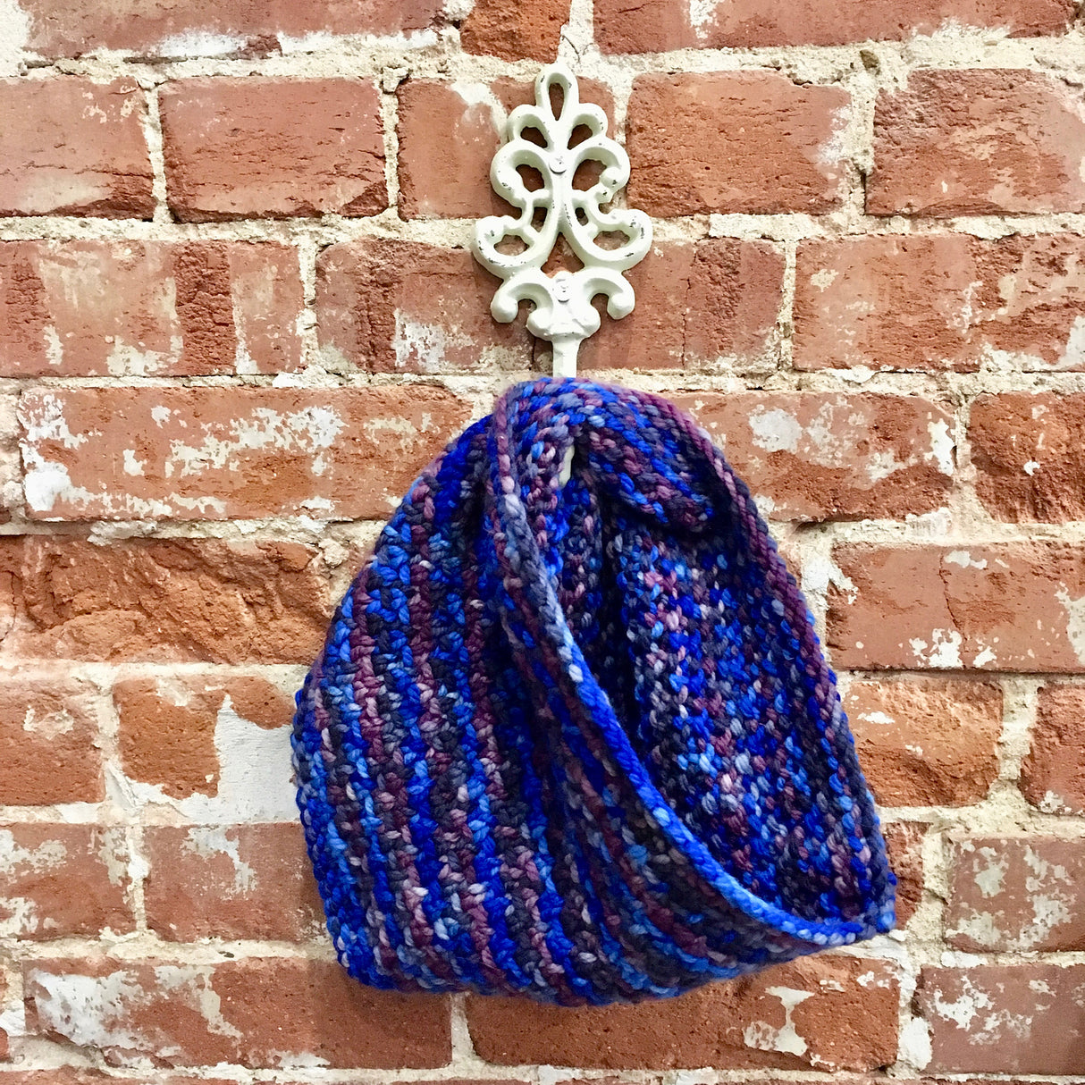 A textured stitch cowl hangs from a white metal hook against a white and brick wall.