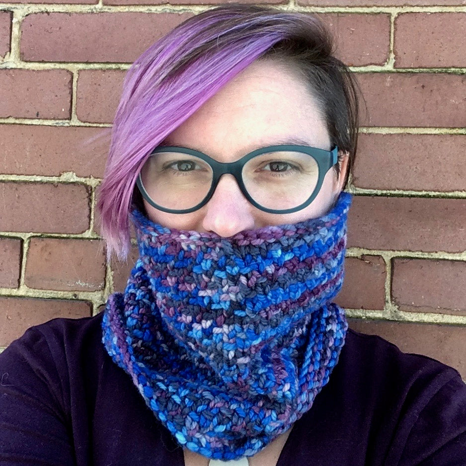A purply-rose haired, light skinned person models a textured cowl in shades of blue. They are wearing dark glasses and a dark top and stand before a brick wall.