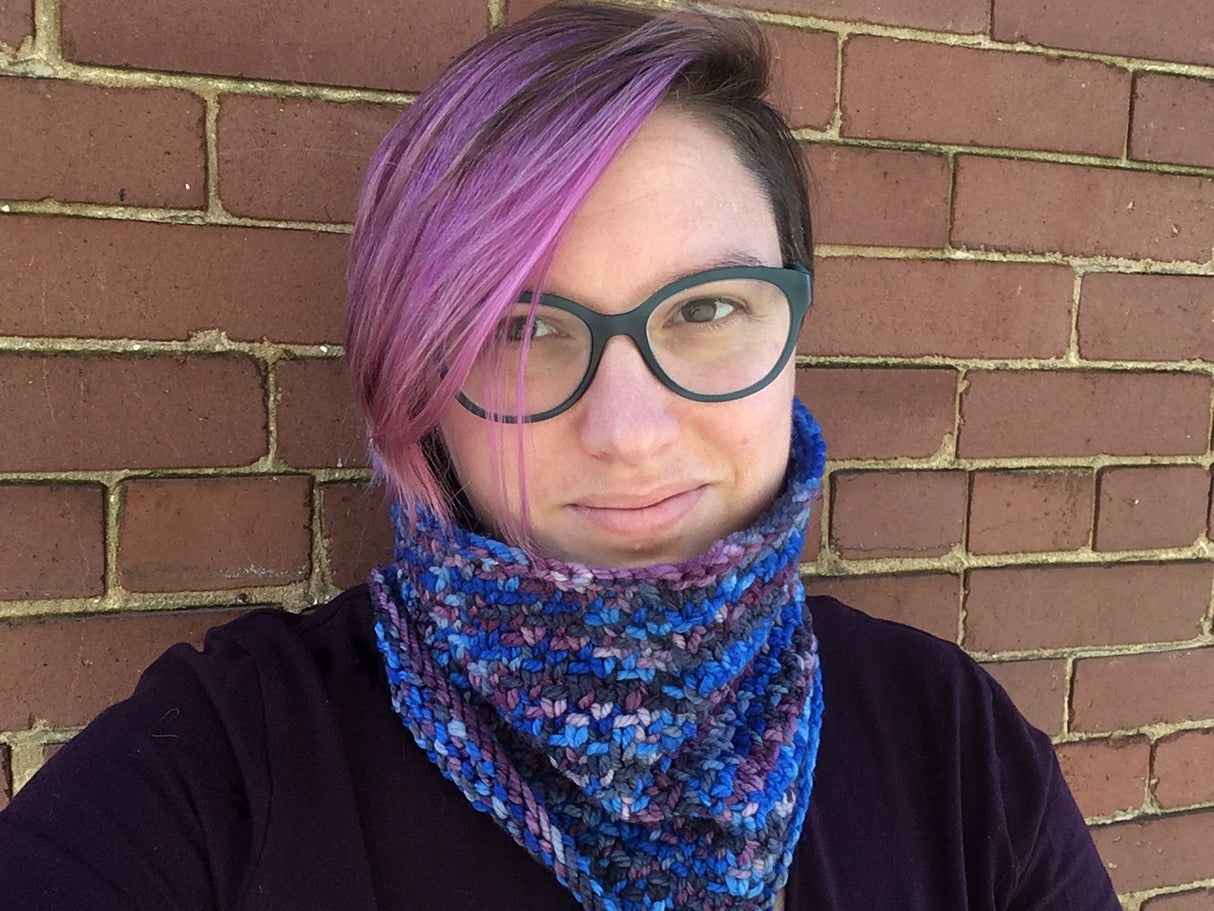 A softly smiling light skinned person stands against a brick wall modeling a textured cowl. Their pinky-purple hair and dark glasses set the yarn off perfectly.