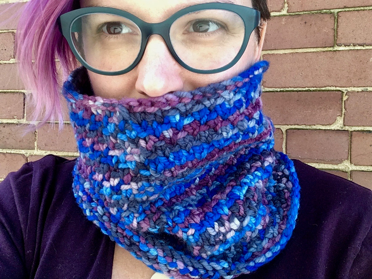 A lovely rose-haired person peeks out fromn behind a richly textured cowl. They are wearing dark glasses and a deep purple top.