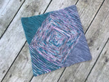 A cowl made of garter triangles in a variety of colors rests on a pale wooden background.