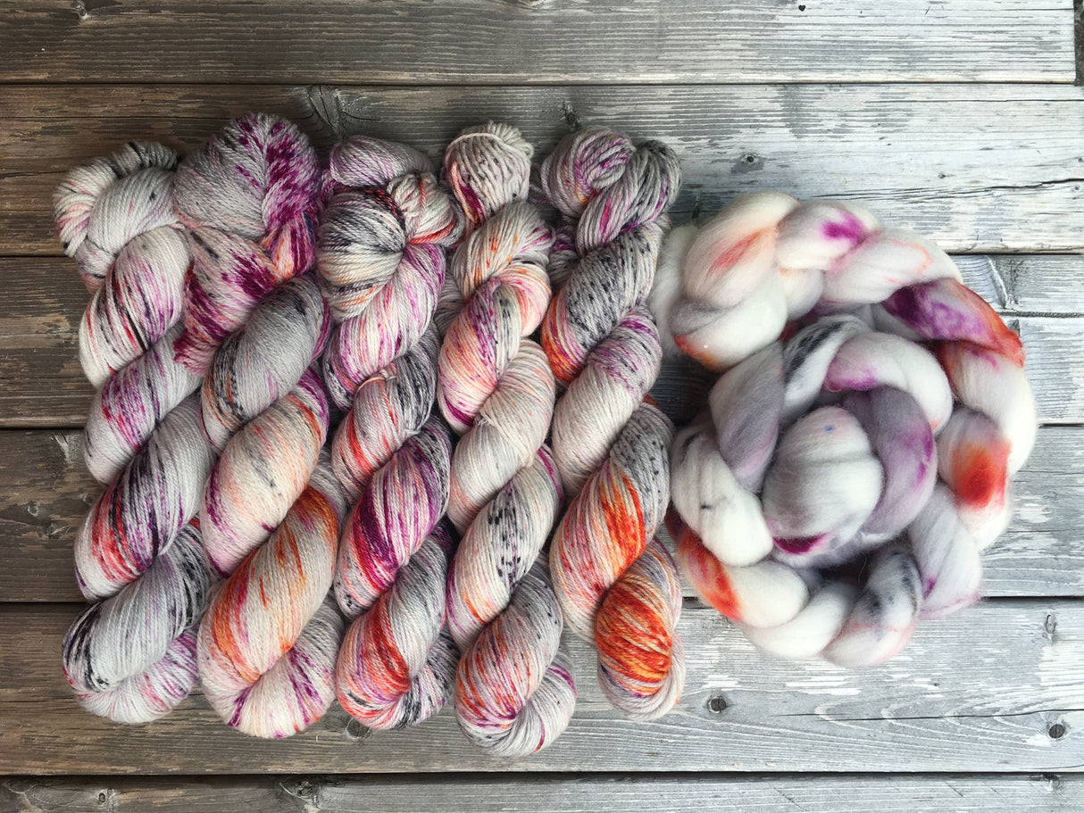 Five vibrant skeins and one fluffy braid of roving rest atop a pale grey wooden background.