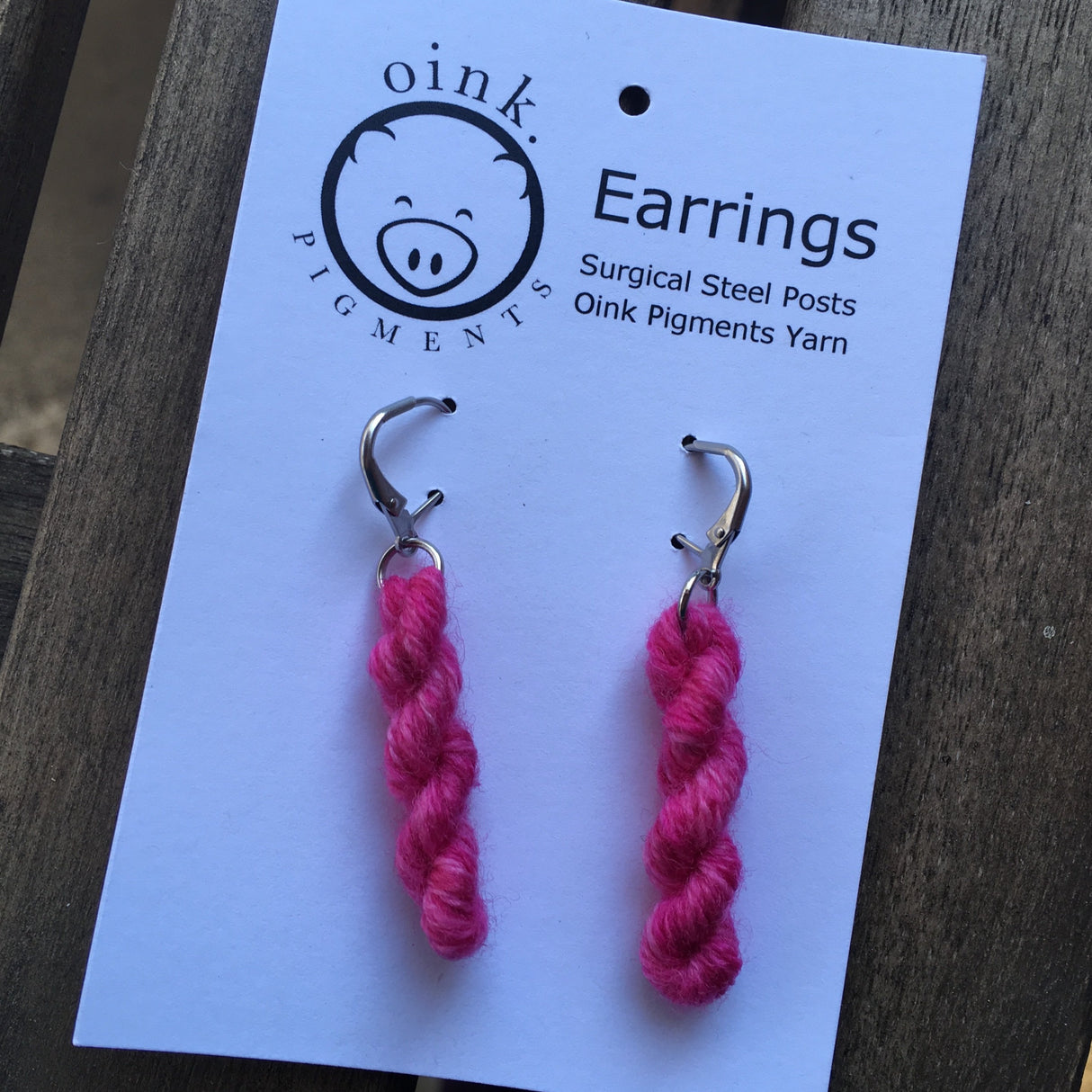 miniature skeins of yarn dangle from steel earring hooks on a white card with the Oink Pigments logo and information at the top.