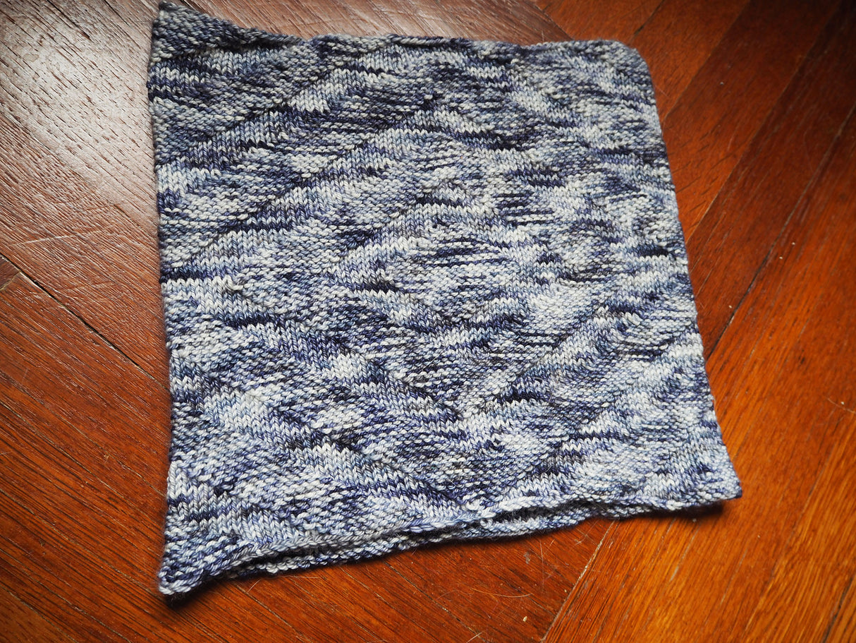 A cowl with a textural herringbone pattern rests against a herringbone patterned wooden floor.