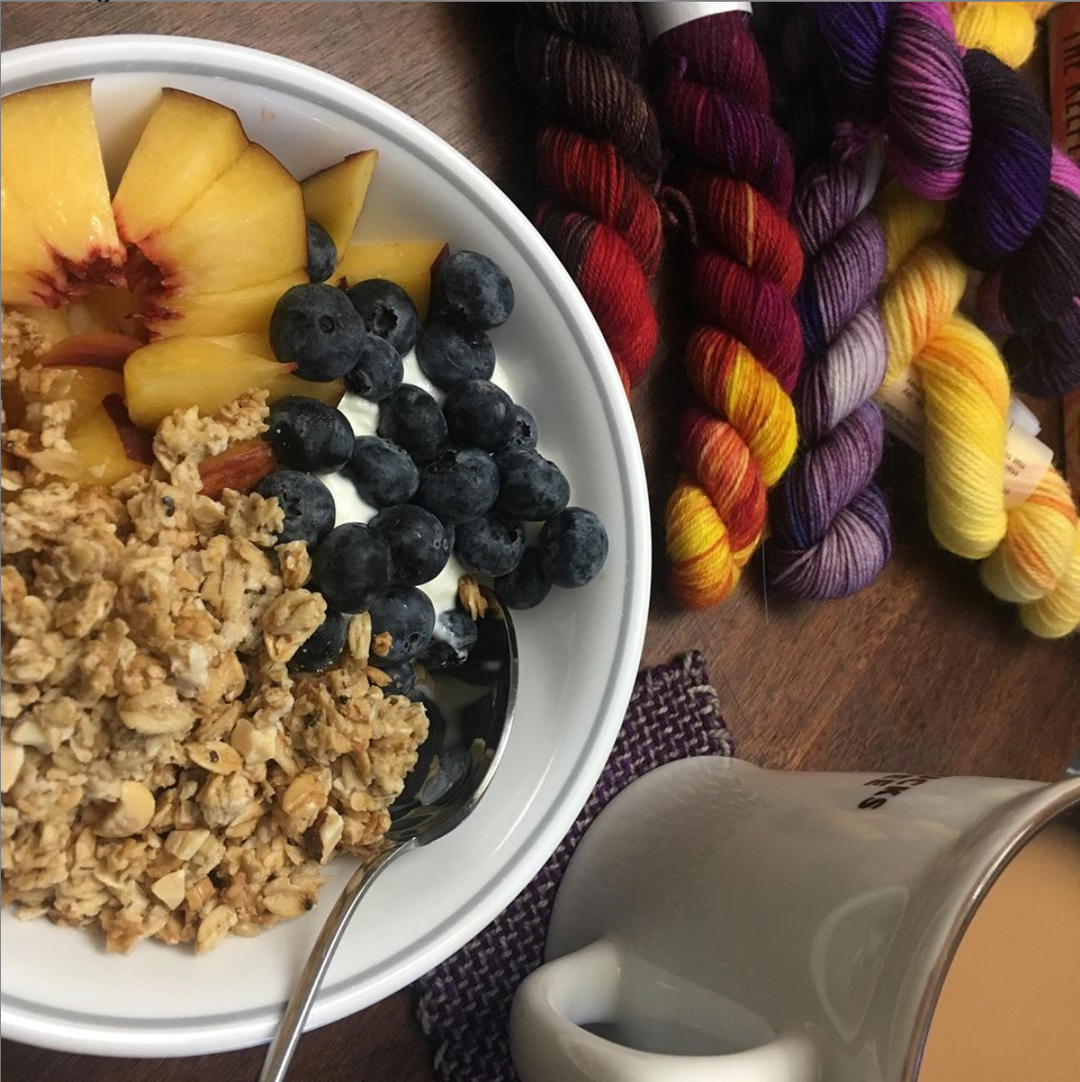 A bowl of granola, blueberries, and nectarine sits beside a cup of coffee. Several skeins of bright yarn rest in the upper right corner.