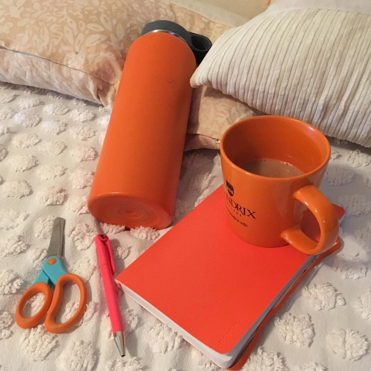Orange! A pair of scissors, a pen, a note book, a huge water bottle and a mug of coffee rest on a cream colored bed spread.