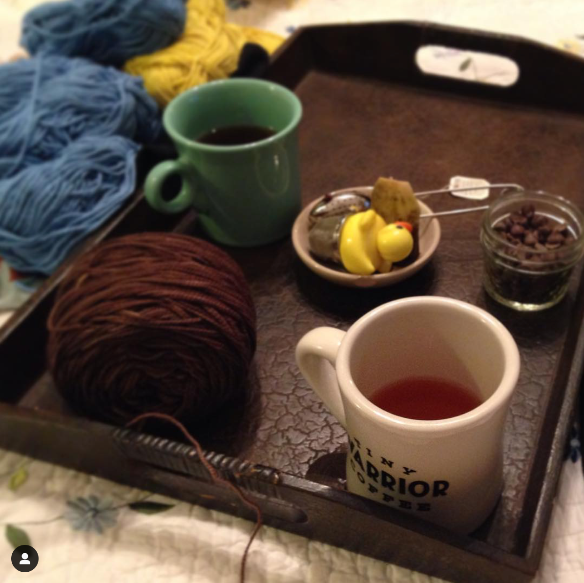 A tray of delight! Two mugs of tea rest on a brown tray with tea accoutrements between them. A cake of chocolaty brown yarn rests on the tray and some soft blue and bright yellow skeins sit to the upper left.