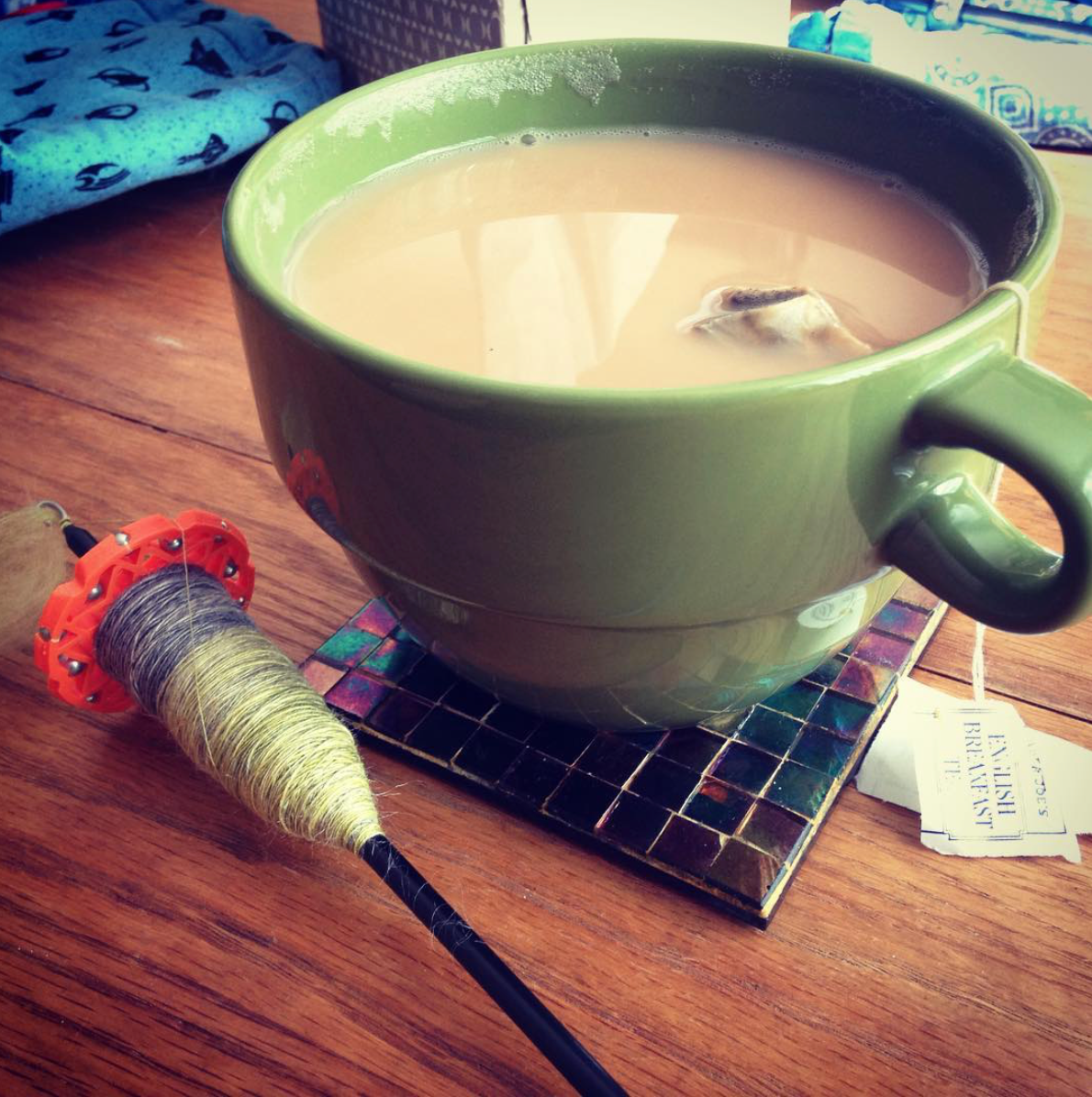 A petite drop spindle rests beside a minty green mug of tea on an oil-slick rainbow coaster.
