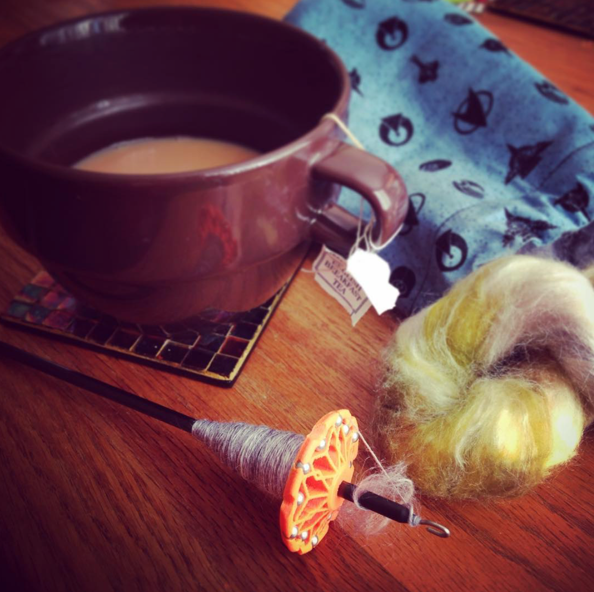 A drop spindle with a twist of colored fluff sit in the foreground. A brown mug of tea and a blue nerdy napkin rest in the background.
