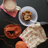 A mug of coffee rests on a knitted swatch in the upper left corner, a milky-white bowl holds a tasty baked good and a project bag with a WIP complete the circle.