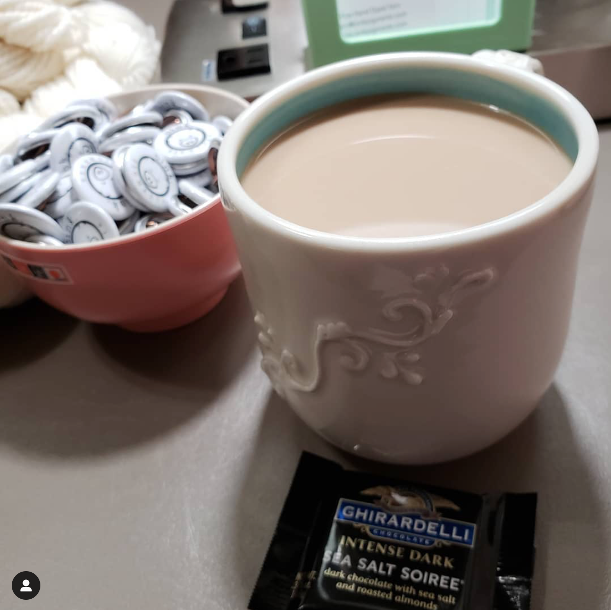 A full mug of wake up beverage awaits with a square of intensely dark chocolate and a bowl of Oink pins around it.