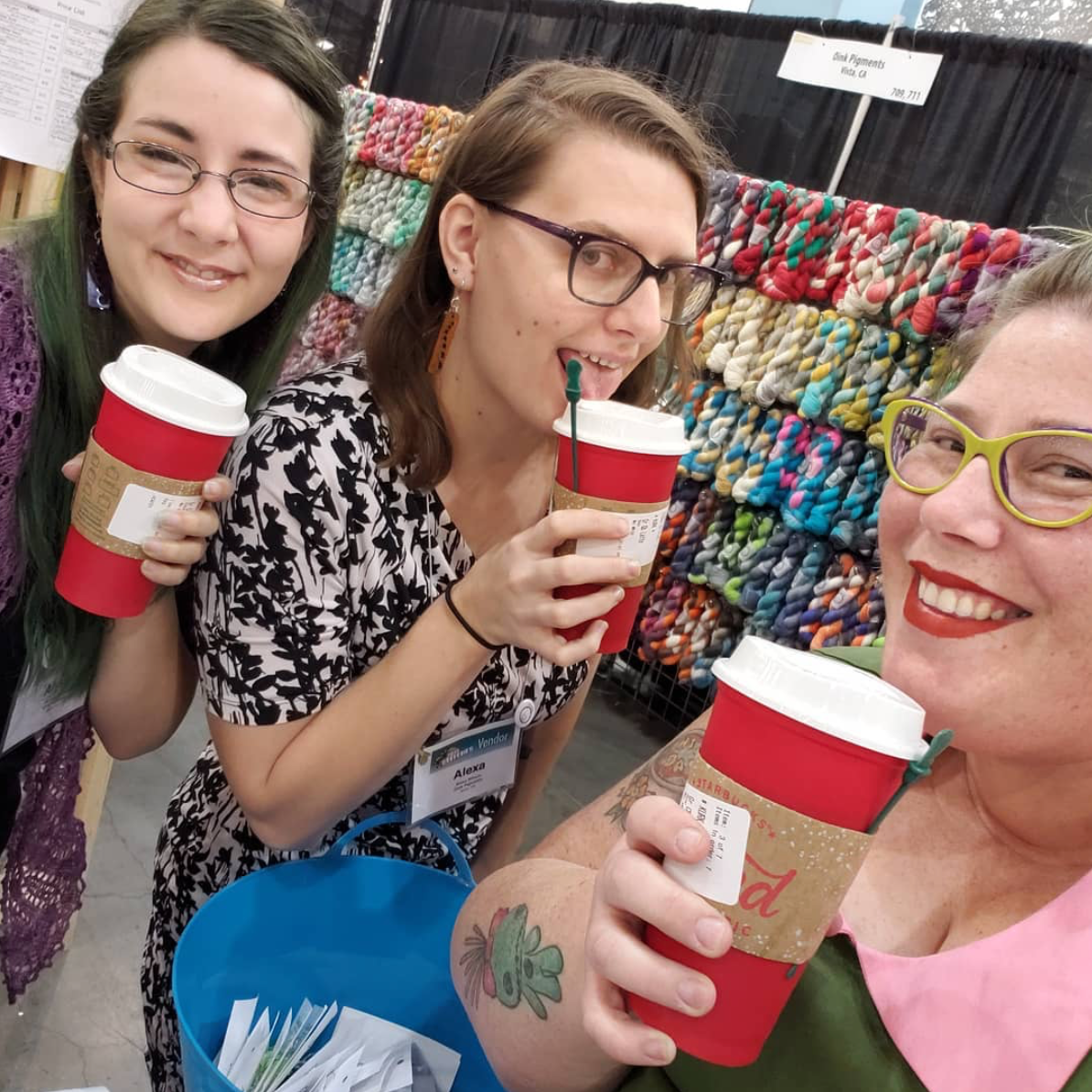 Three smiling faces of Team Oink against a wall of dazzling yarn. Each person is holding a red coffee cup.