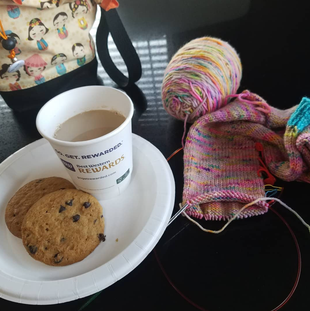 A picture perfect pause; two cookies and a cup of coffee rest on a white paper plate surrounded by a colorful project bag and a sock in progress.