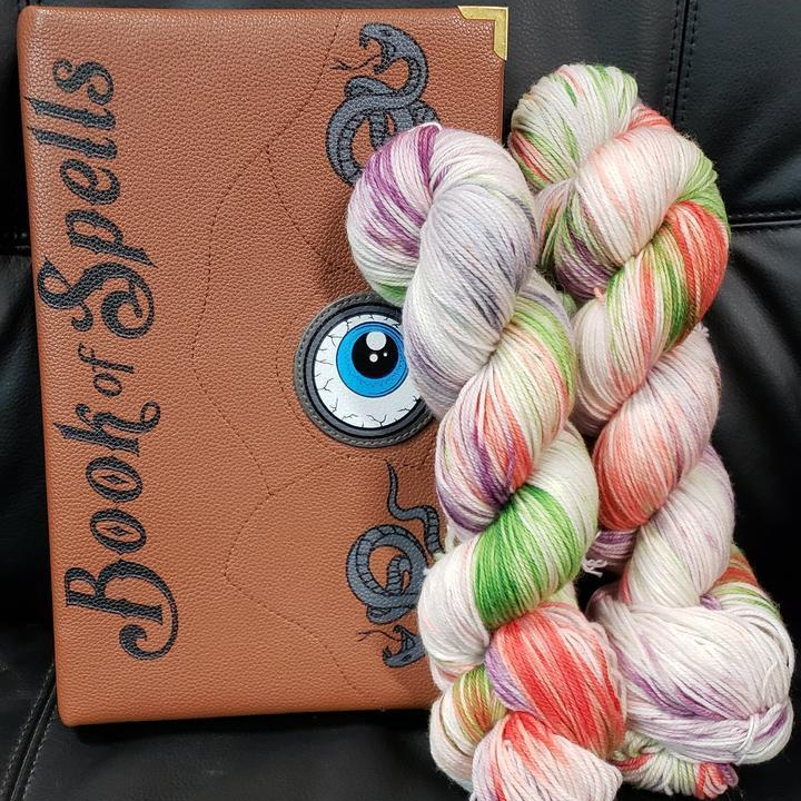 Two skeins of yarn in the "A Pox, a Pox, a Pox!" colorway rest against a brown Book of Spells with a blue eye peering up at the yarn. The background is black.
