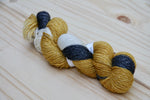 A golden, cream, and black skein of yarn with a subtle glimmer rests on a pale wooden background. 