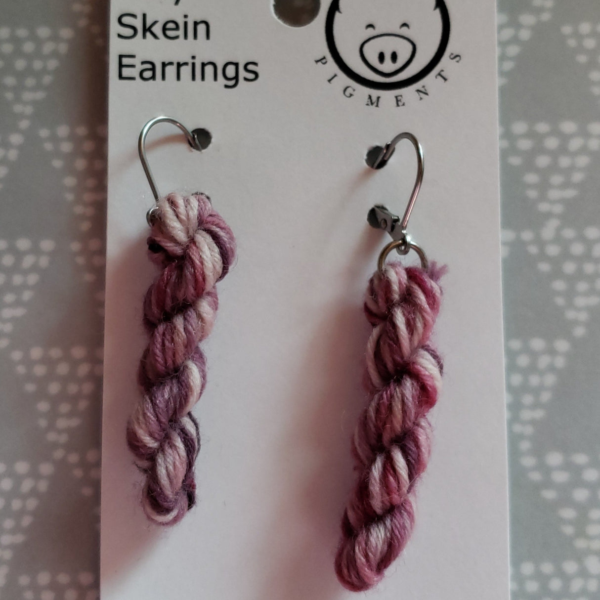 miniature skeins of yarn dangle from steel earring hooks on a white card with the Oink Pigments logo and information at the top.