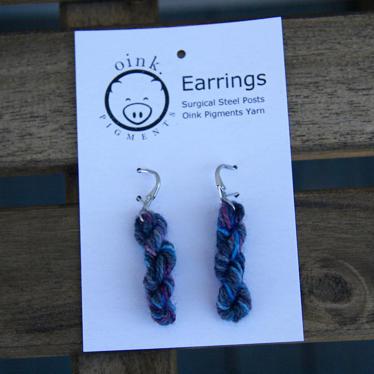 miniature skeins of yarn dangle from steel earring hooks on a white card with the Oink Pigments logo and information at the top.