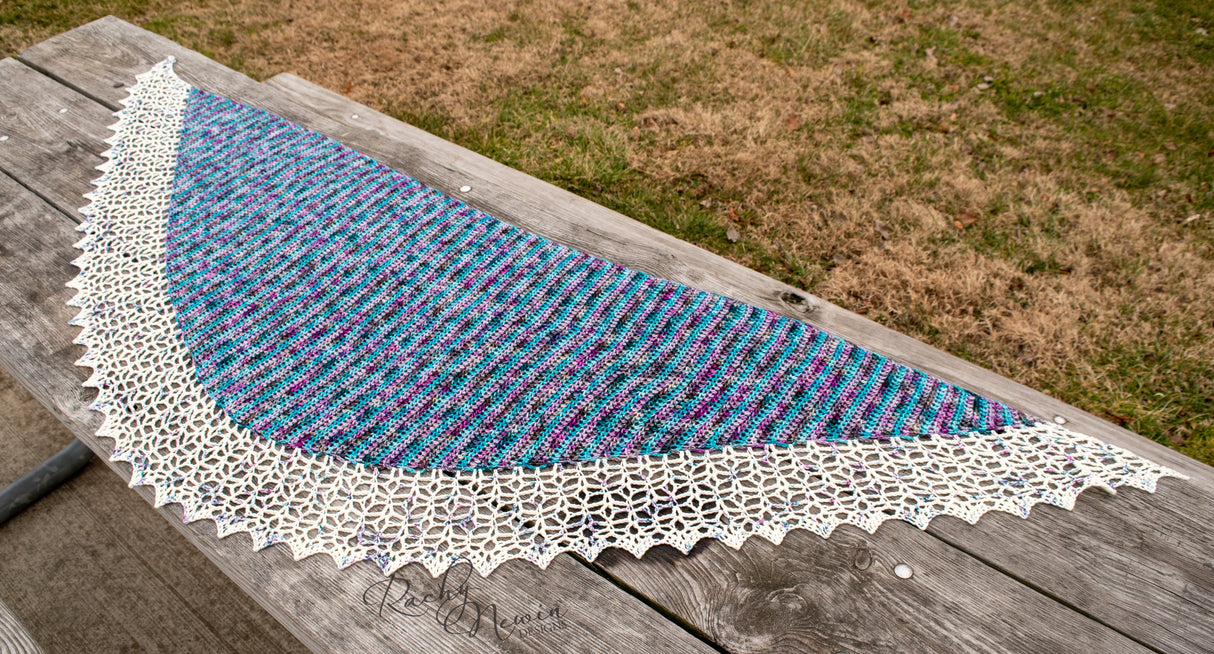 An image of the wingspan and general shape of the shawl as it rests on a well loved picnic table. Brown-green grass is visible in the upper right corner of the frame.