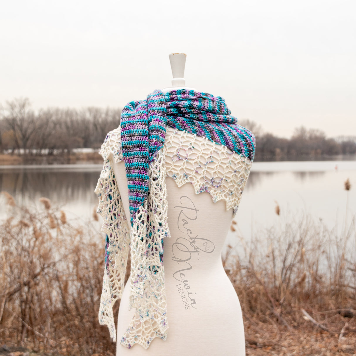 An image of the Bonfire Shawl wrapped around the shoulders of a white mannequin against a brown & silver lakeside background.
