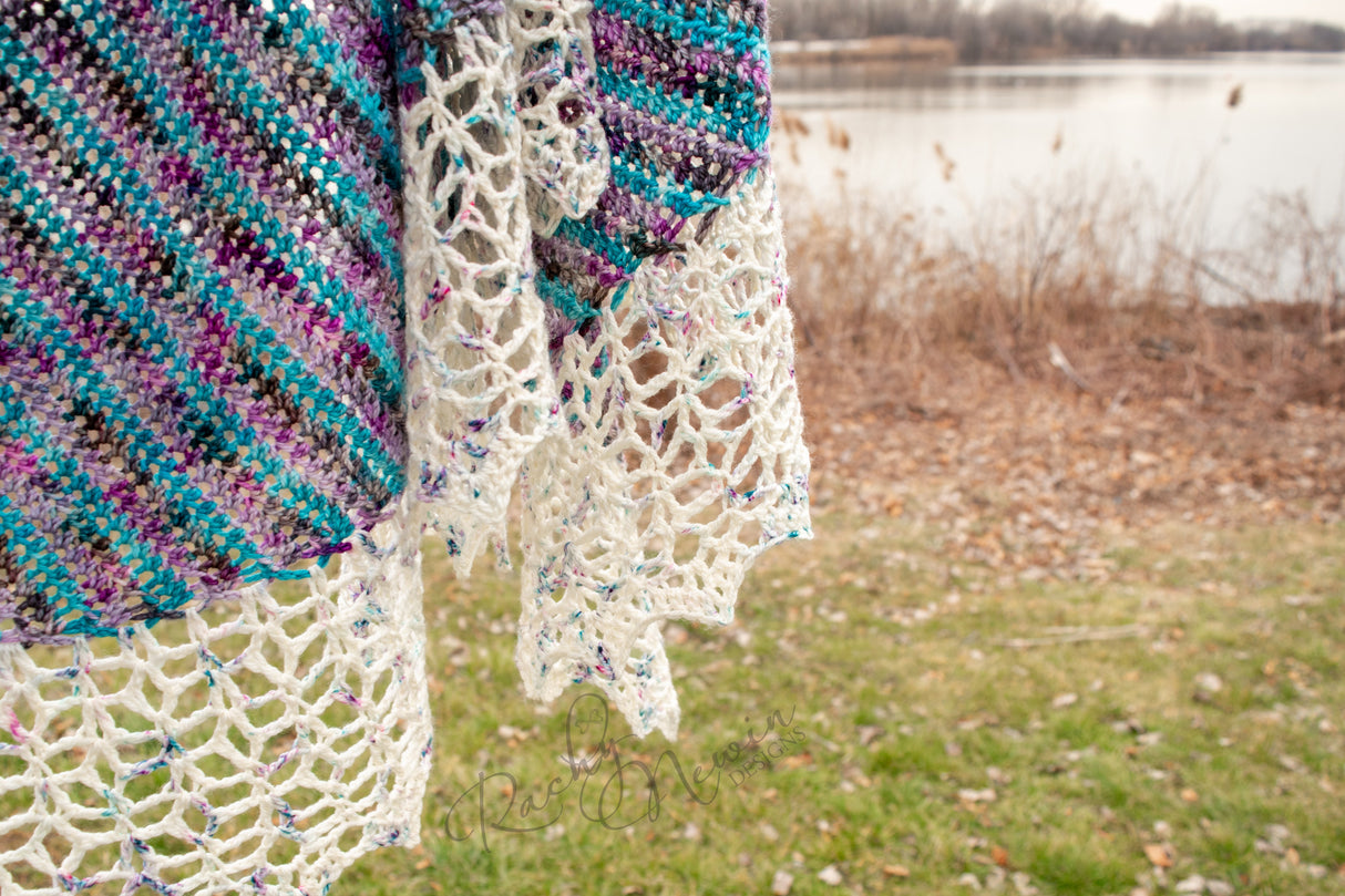 A detail image showing the slight open crochet pattern of the main shawl and the delicate lace edge. There is green and brown grass and a silvery lake in the background.