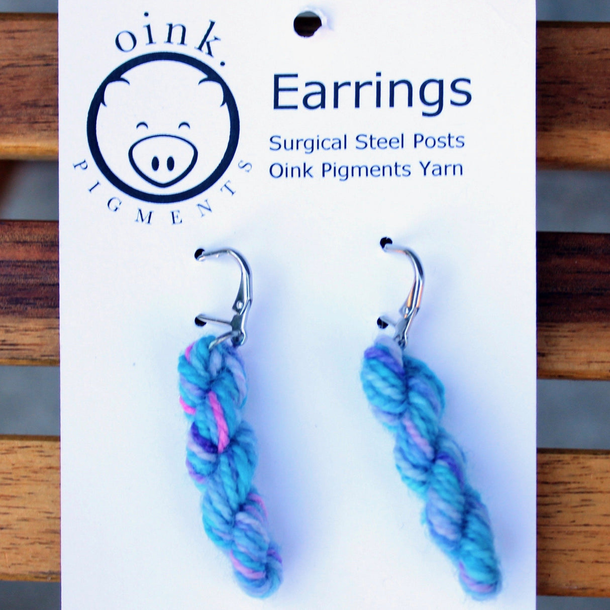 miniature skeins of yarn dangle from steel earring hooks on a white card with the Oink Pigments logo and information at the top.
