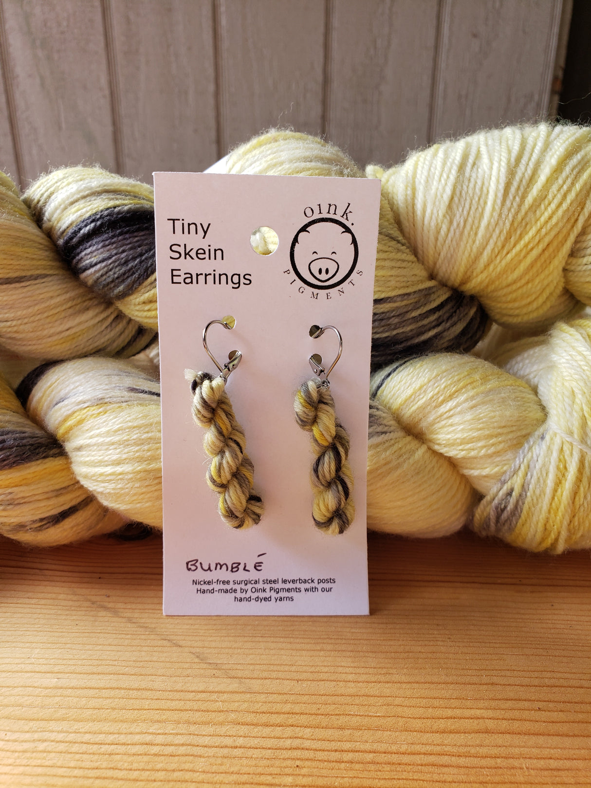 miniature skeins of yarn dangle from steel earring hooks on a white card with the Oink Pigments logo and information at the top. The card is resting against two skeins of yarn.