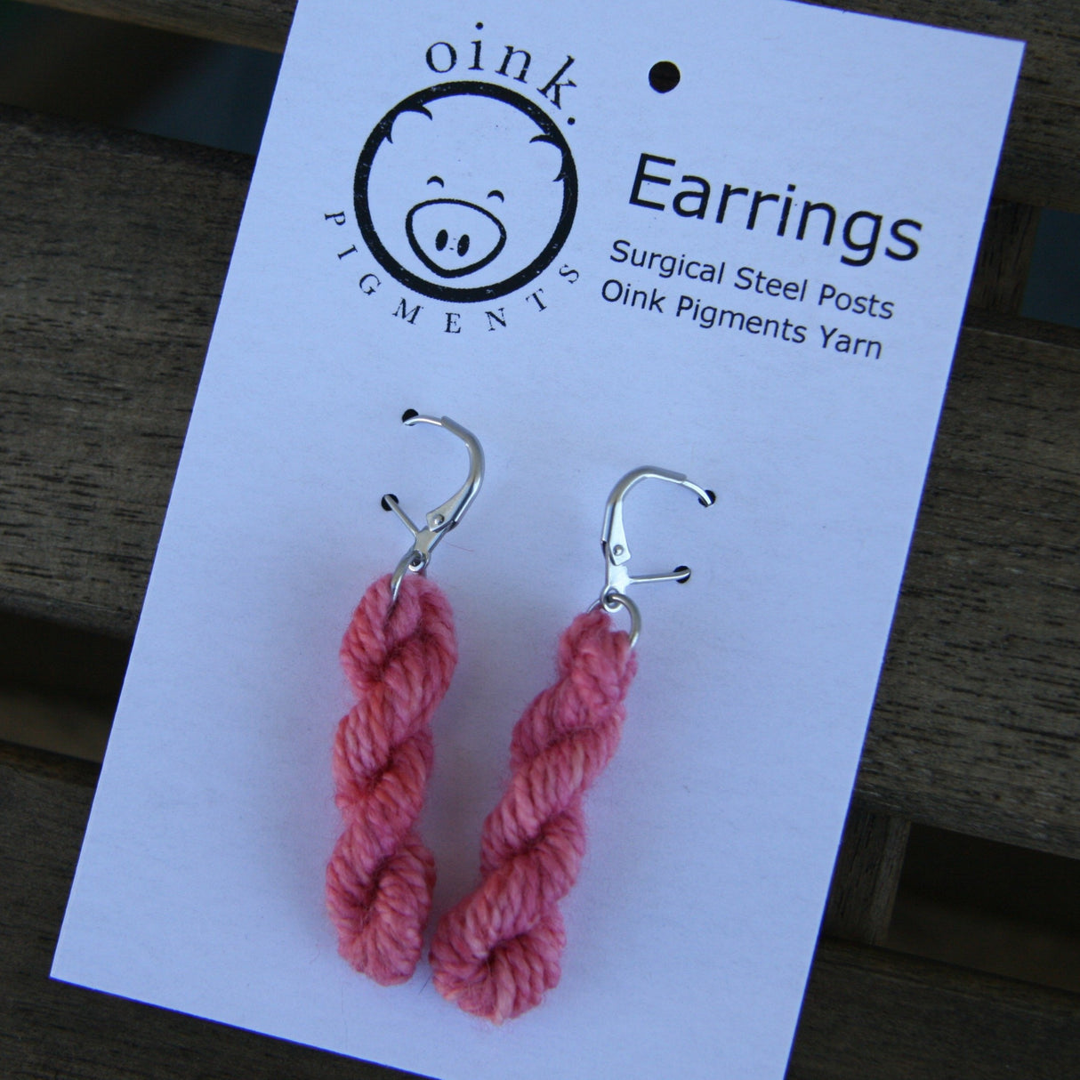 miniature skeins of yarn dangle from steel earring hooks on a white card with the Oink Pigments logo and information at the top.