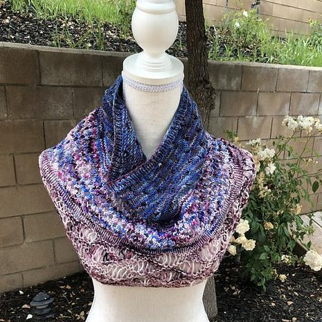 A textural cowl in shades of purple, rose, and cream rests on the neck of a mannequin form. There is a grey brick wall and some greenery in the background.