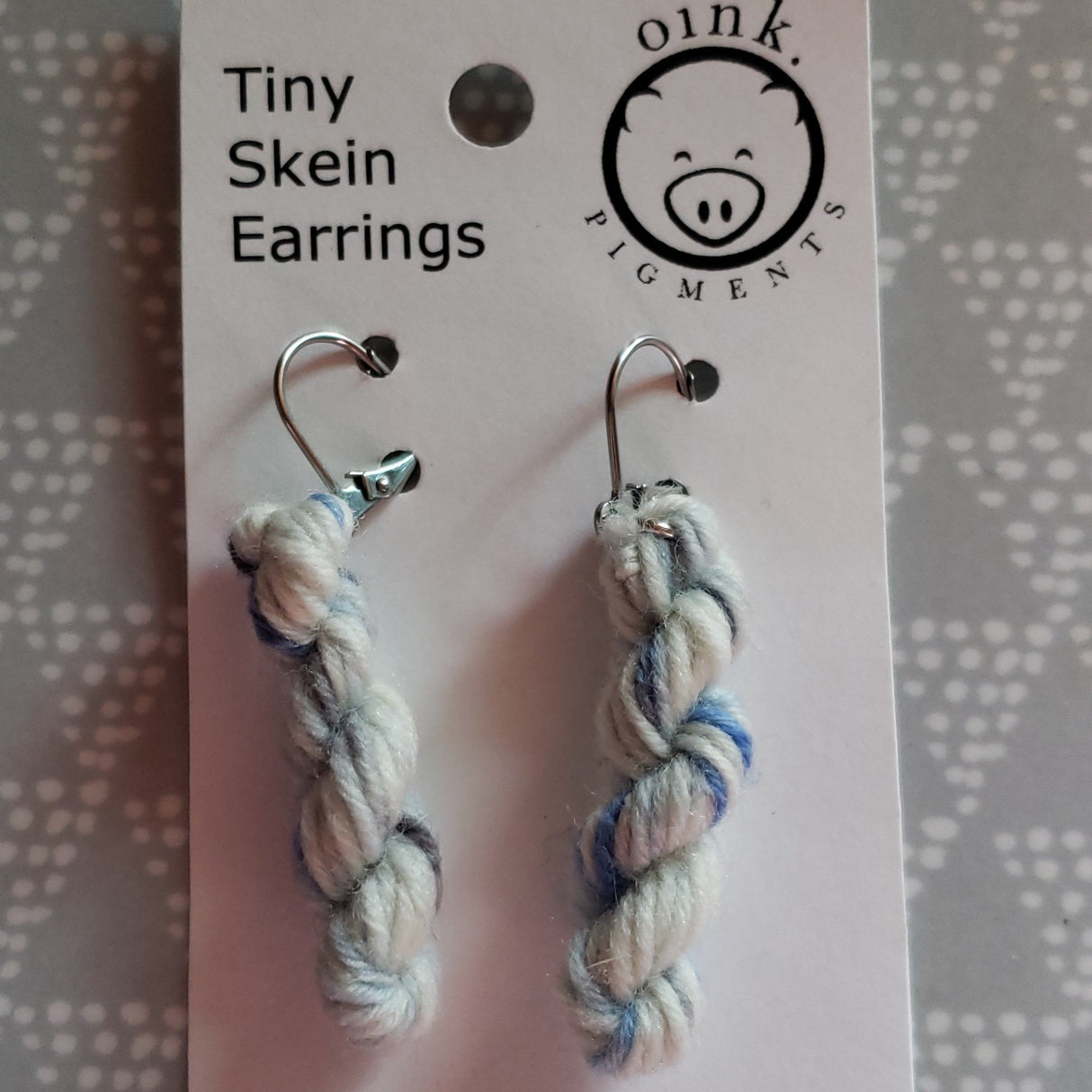 miniature skeins of yarn dangle from steel earring hooks on a white card with the Oink Pigments logo and information at the top.