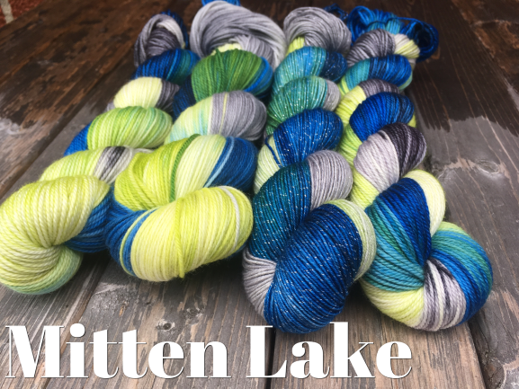 four skeins of yarn sit on a wooden background, with tones of deep blue, lime, grey, and chartreuse. White text at the bottom left reads "mitten lake"