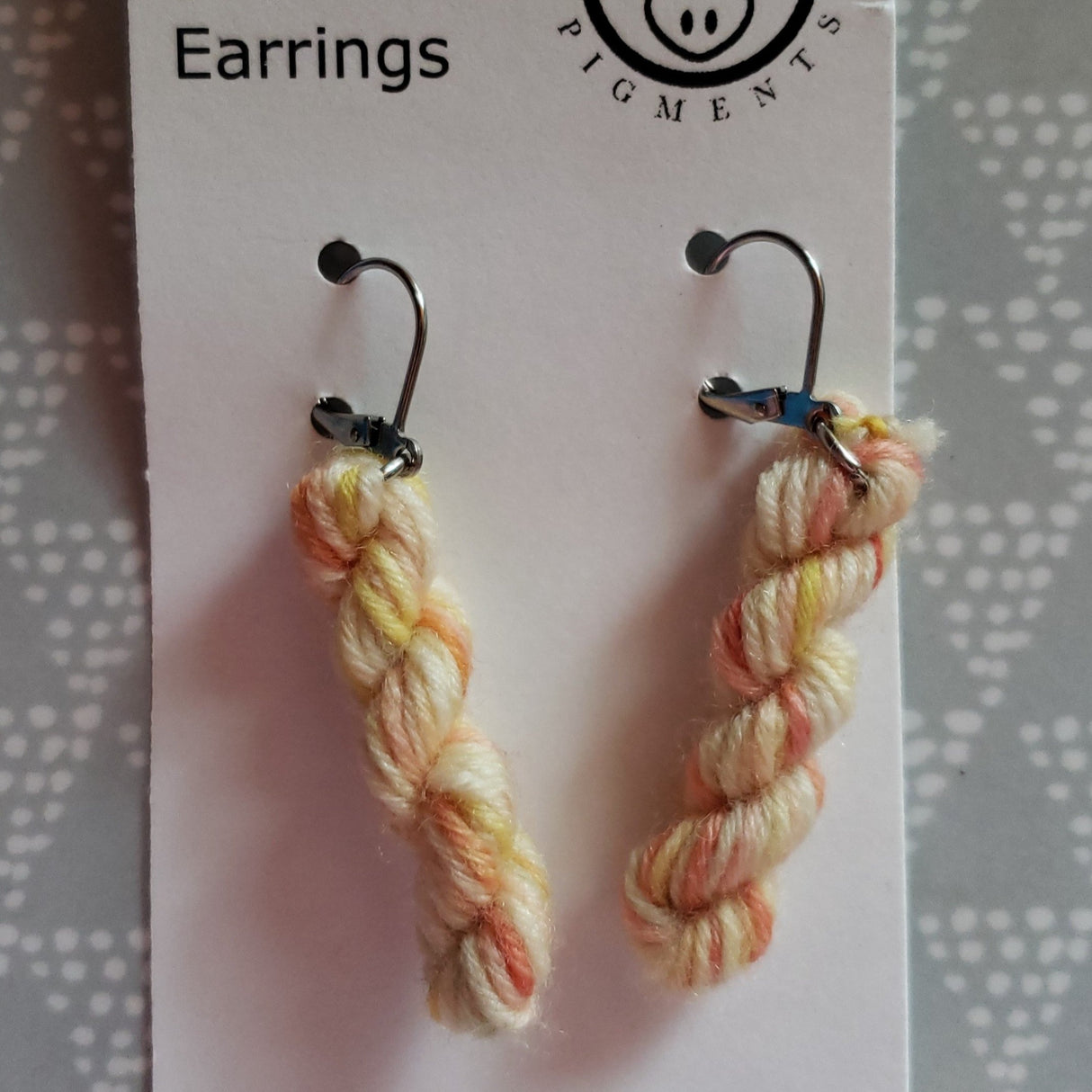 miniature skeins of yarn dangle from steel earring hooks on a white card with the Oink Pigments logo and information at the top.