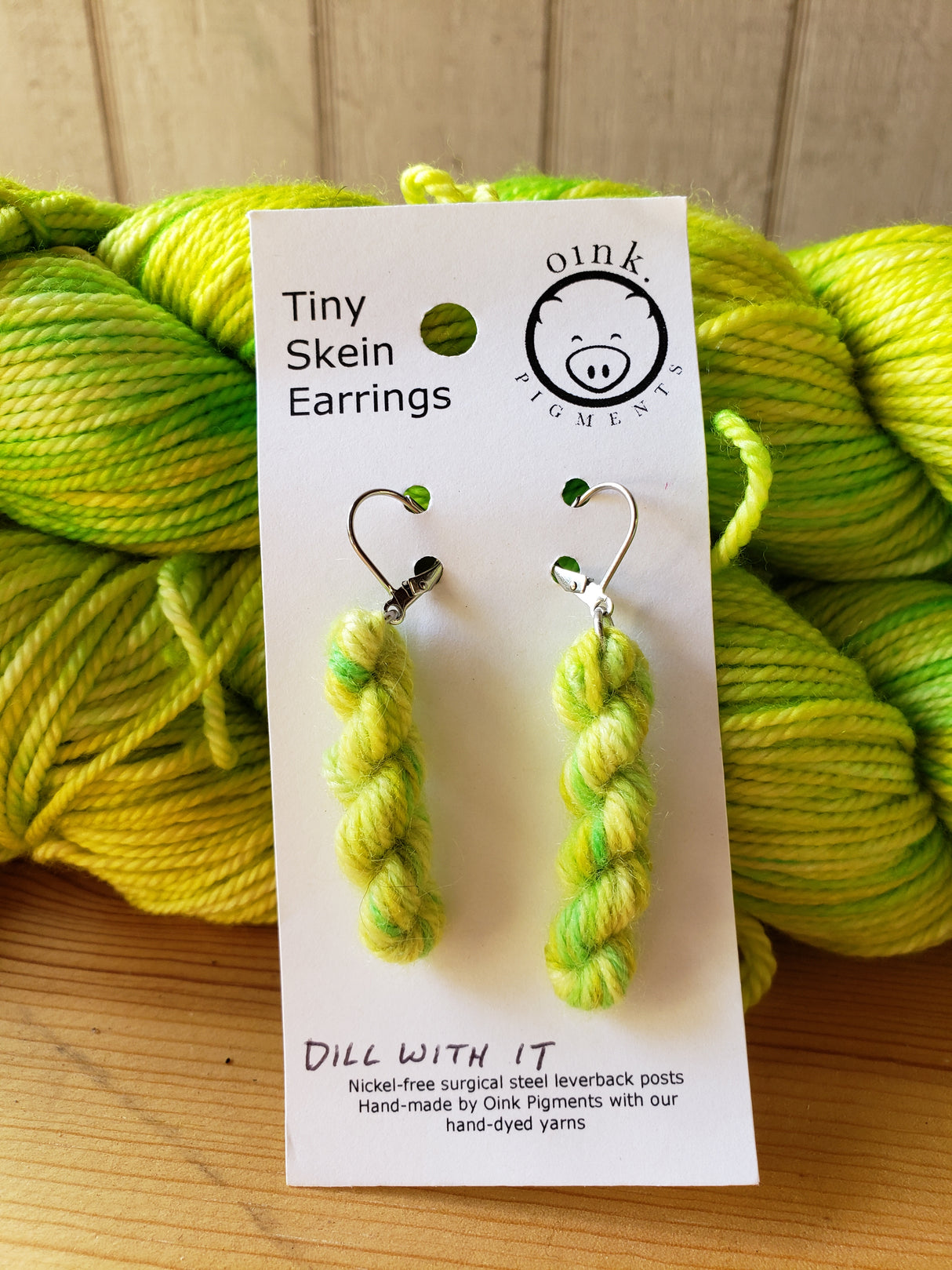 miniature skein earrings rest on a white card with the Oink Pigments logo and text at the top of the card. The card rests on a stacked pair of skeins of yarn.