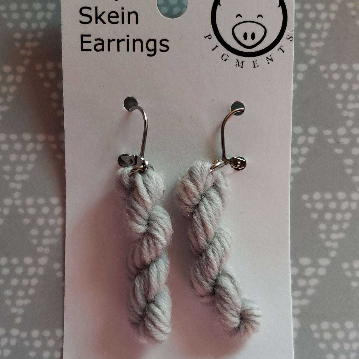 miniature skeins of yarn dangle from steel earring hooks on a white card with the Oink Pigments logo and information at the top.