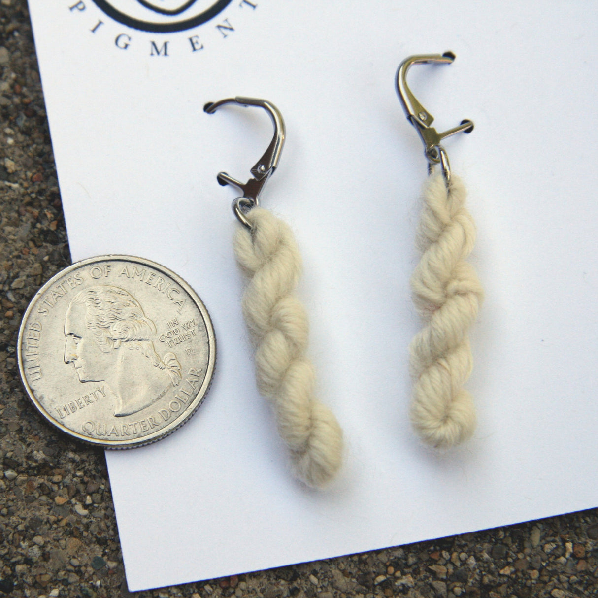 miniature skeins of yarn dangle from steel earring hooks on a white card with the Oink Pigments logo and information at the top. A quarter rests to the left of frame for size reference.
