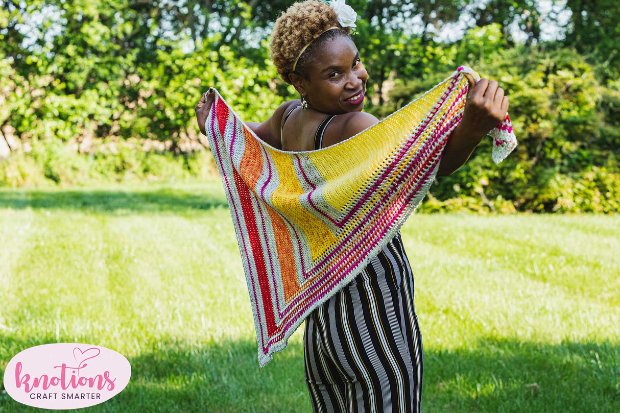 A sweetly smiling medium skinned person stands in a grassy field holding a bright and colorful shawl behind them. There is a small pink label in the bottom left corner of the frame.