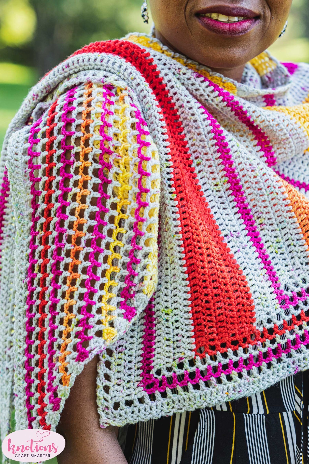 A close up shot of a crocheted shawl. There is a soft smile on a dark skinned person's face in the upper right corner.